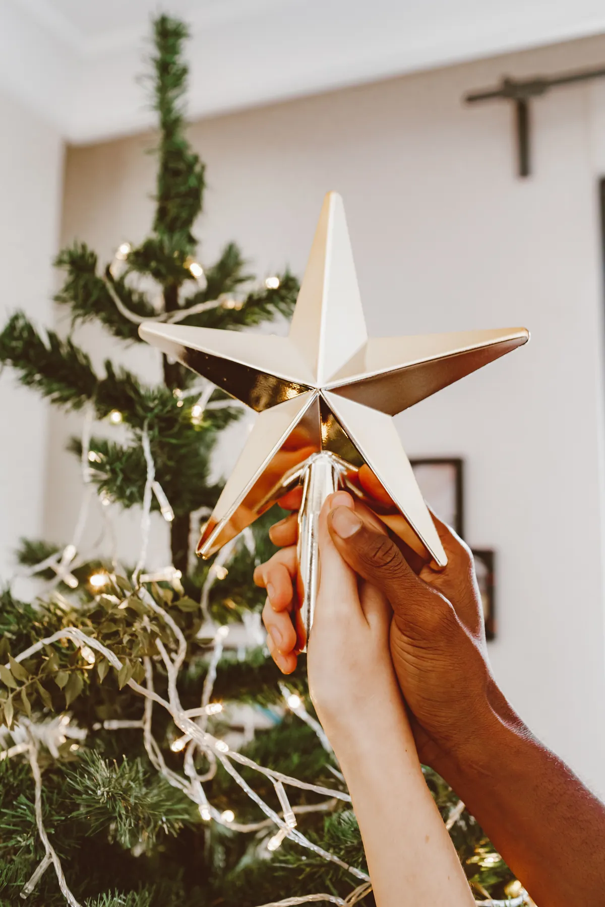 Two hands of different ethnicities place a star atop a Christmas tree indoors.