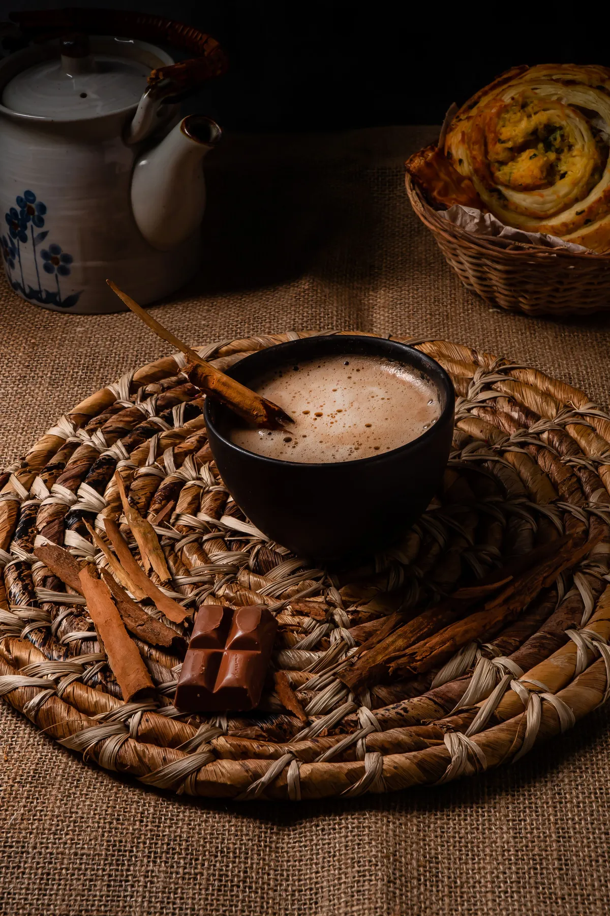 Warm hot chocolate with cinnamon sticks and pastries, styled on a woven placemat.