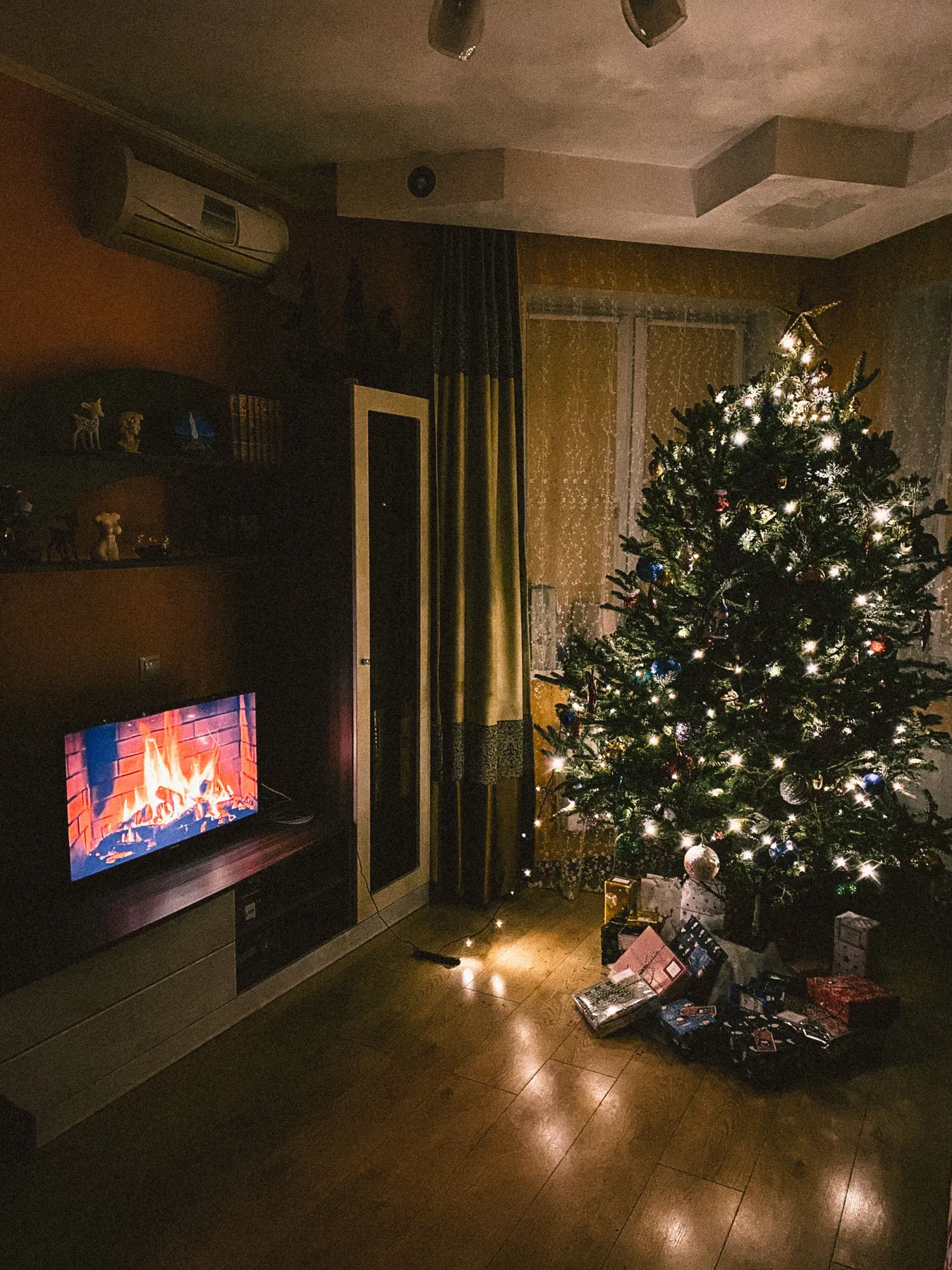 Warm indoor Christmas scene with tree, lights, gifts, and cozy fireplace on TV.