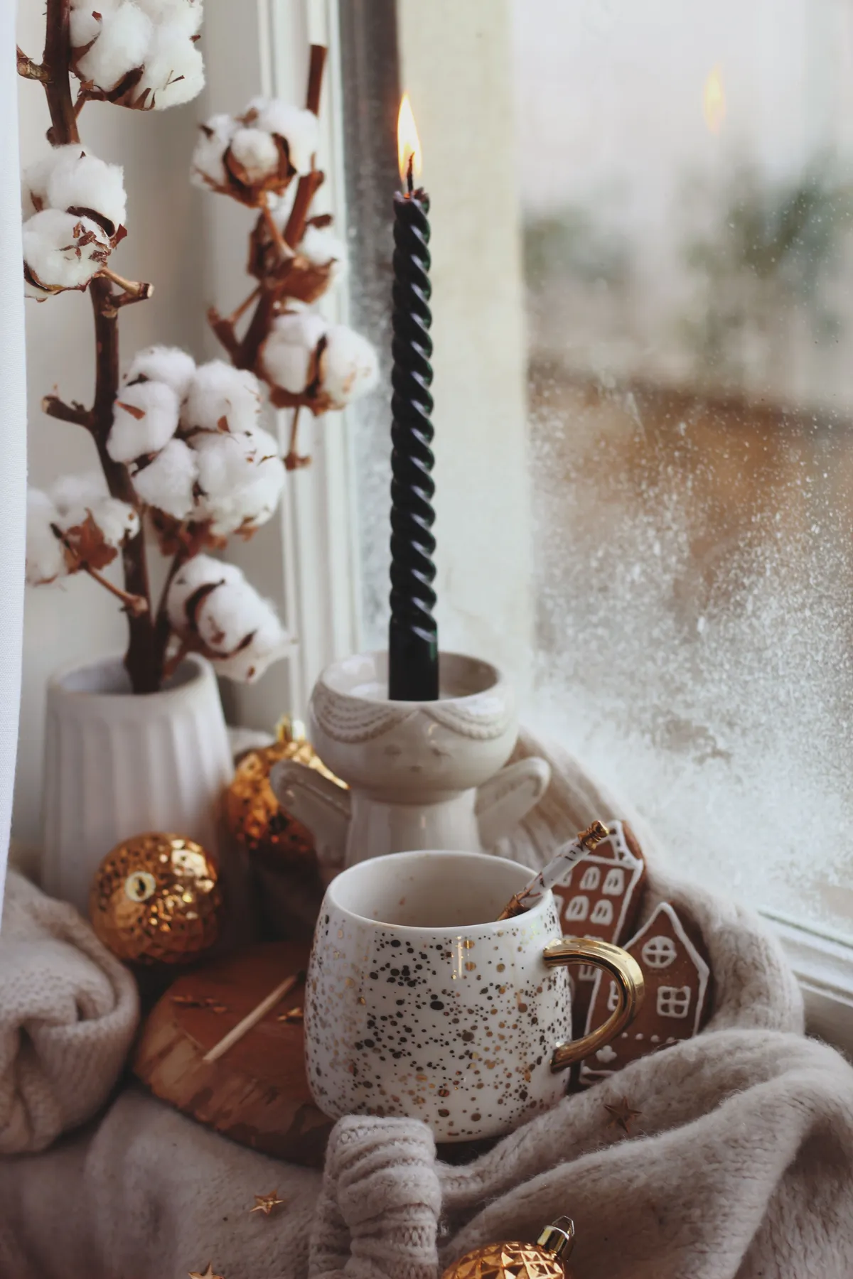 Warm winter decor with cotton branches, candle, and hot drink near a frosty window.