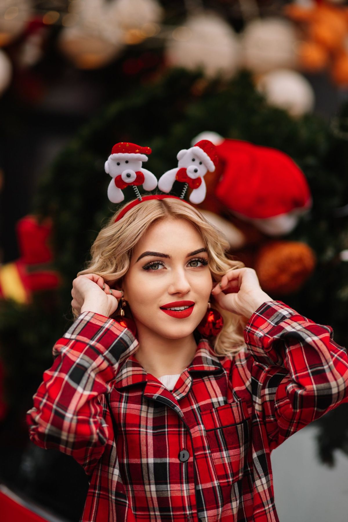 Woman in red plaid pajamas and Santa headband celebrating Christmas indoors with a festive background.